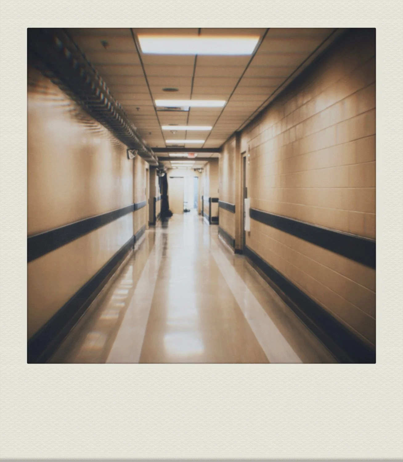 Long hospital corridor with beige walls, black horizontal stripes, fluorescent ceiling lights, and a person standing near a door at the end.