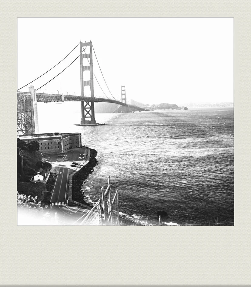 Black and white photo of the Golden Gate Bridge over water with a historic fortress and parking lot in the foreground.