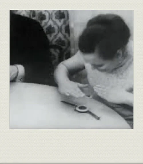 A woman appears to be drawing or writing on a person's arm as they are seated at a table with a magnifying glass nearby.
