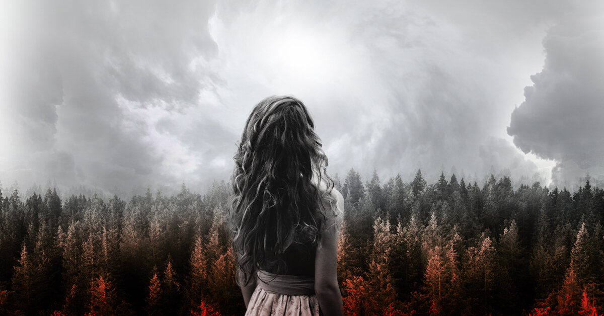 Back view of a girl with long wavy hair standing in front of a forest under a cloudy sky.