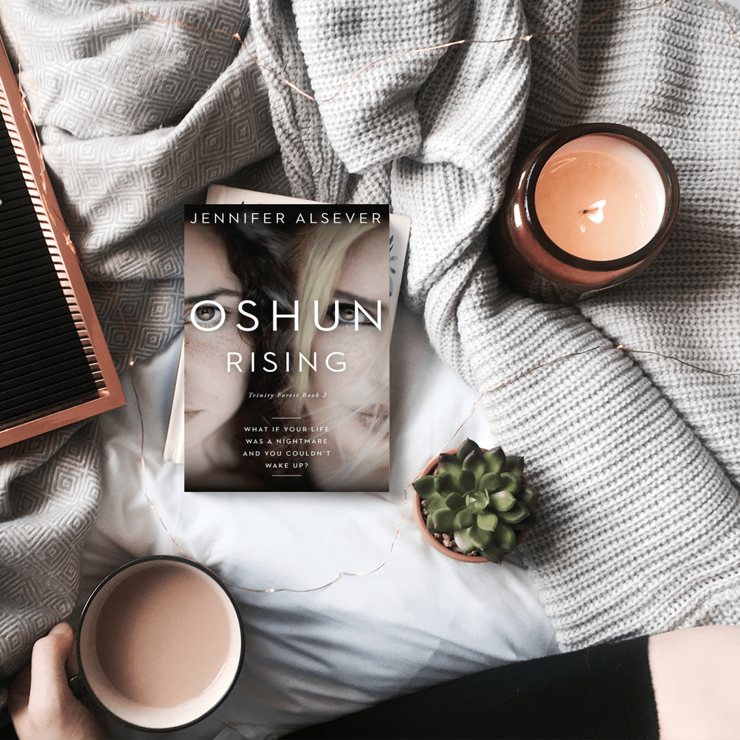 A cozy bedroom scene with a book titled 'Oshun Rising' by Jennifer Alsever, two coffee mugs filled with a light brown beverage, a small potted succulent, a pink hairbrush, a grey patterned blanket, a grey textured blanket, a lit candle in a brown jar, and a string of fairy lights.