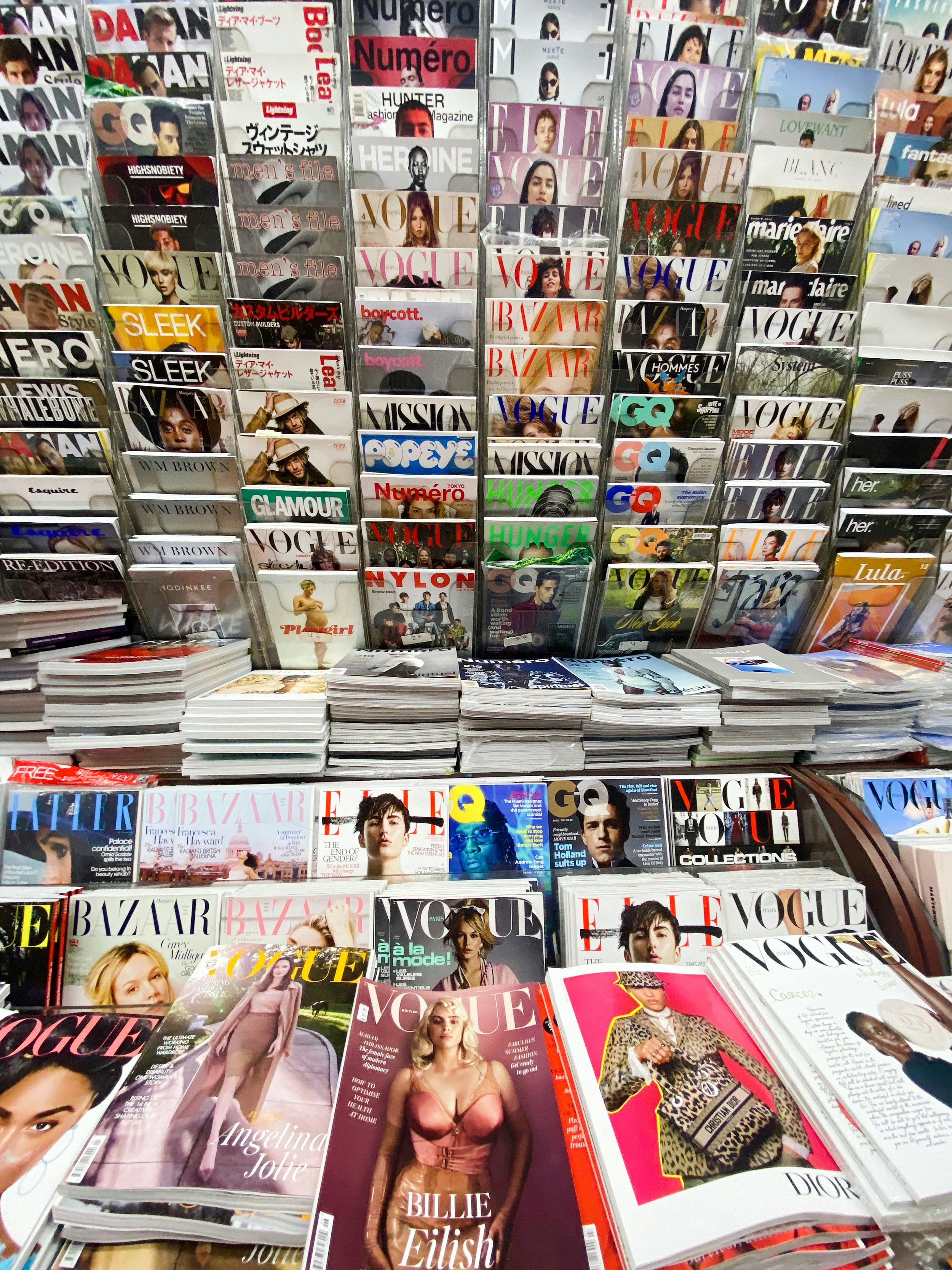 Display of various fashion magazines, including Vogue, Harper's Bazaar, Elle, and Marie Claire, arranged on racks and tables at a magazine store.