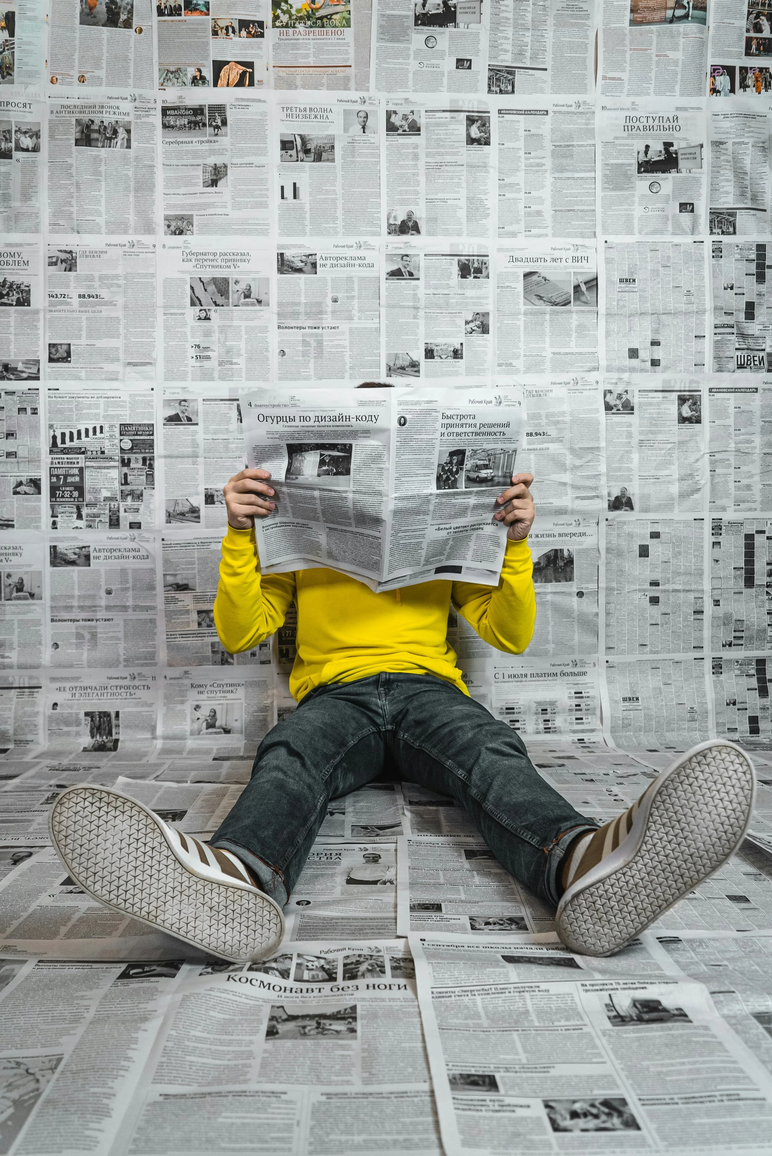 Person sitting on the floor against a wall covered with newspapers, reading a newspaper. The person is wearing a yellow long-sleeve shirt, black jeans, and white sneakers. The background and floor are entirely covered with newspapers.