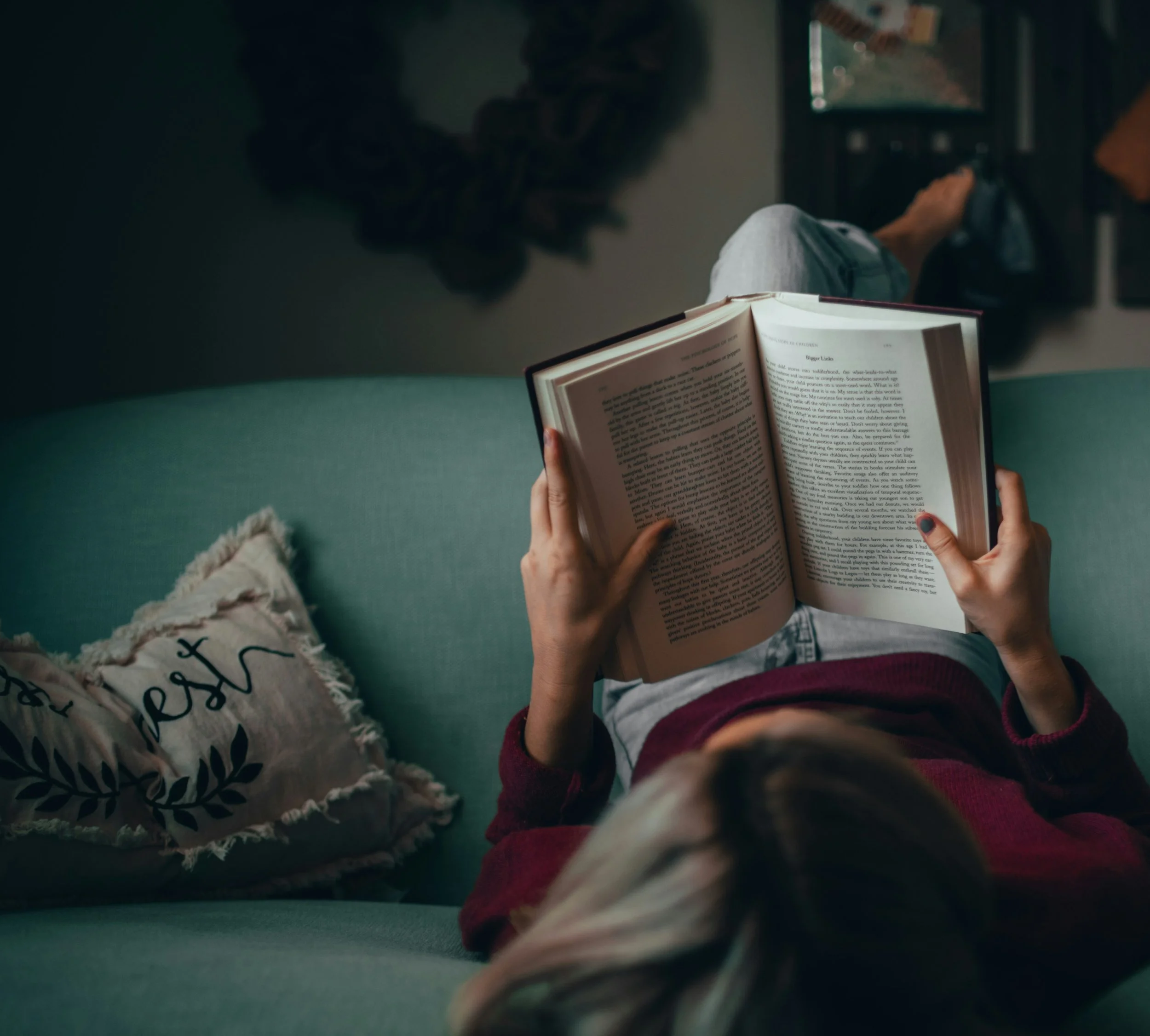 Person lying on a green couch reading an open book, with a decorative pillow nearby and a blurry background.