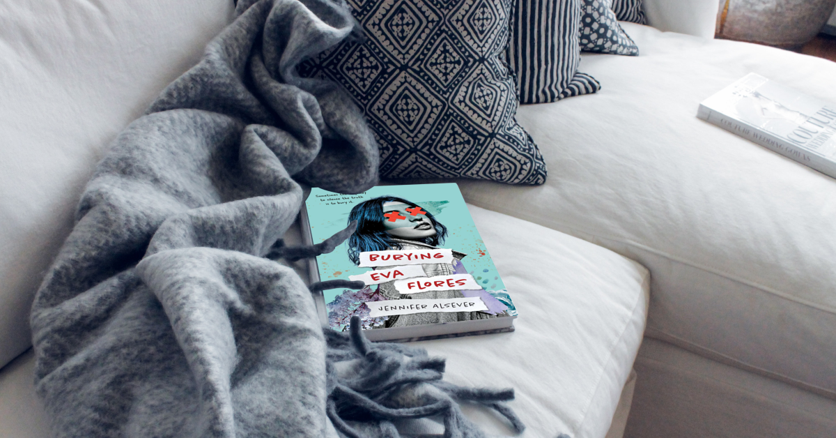 A paperback book titled 'Burying Eva Flores' by Jennifer Alsever resting on a white couch, partially covered by a soft gray blanket with a textured blue and white pillow nearby. Another book is visible on a side table.