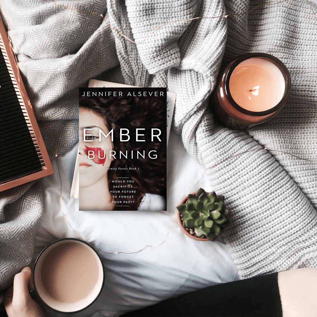 A cozy setup with a book titled 'Ember Burning' by Jennifer Alsever on a bed surrounded by gray blankets, a small potted succulent, two candles, and a cup of tea or coffee.