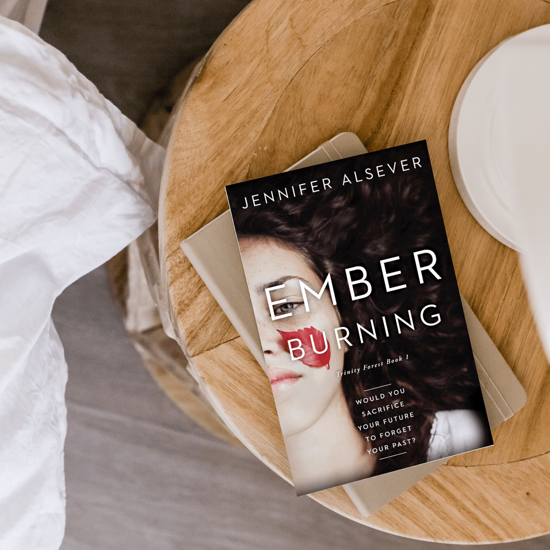A book titled 'Ember Burning' by Jennifer Alsever resting on a beige notebook on a round wooden table, next to a white plate.