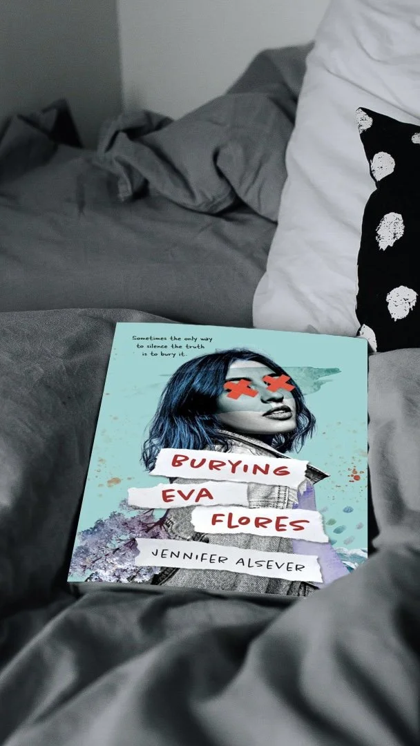 A book titled 'Burying Eva Flores' by Jennifer Alsever lying on a gray bed with gray and white pillows.