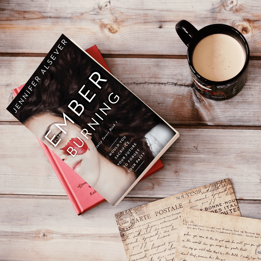 A book titled 'Ember Burning' by Jennifer Alsever lies on a wooden surface, with a cup of coffee or tea nearby, and some vintage postcards.