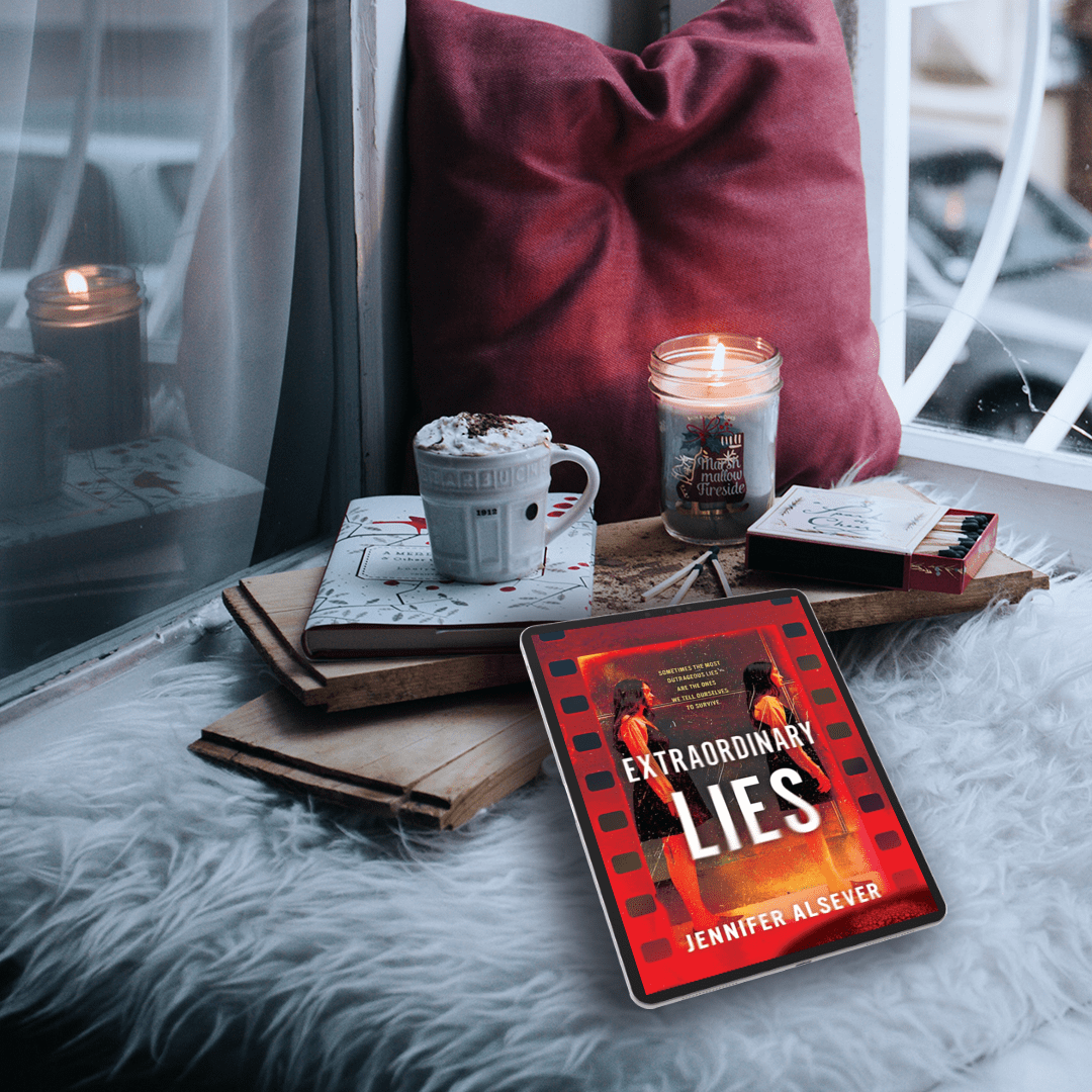A cozy scene with a tablet displaying the book cover "Extraordinary Lies" by Jennifer Alsever, a cup of hot chocolate with whipped cream, a lit candle, a pack of matches, and a box of matches on a wooden tray. The setup includes a fluffy white surface, a red pillow, and a window in the background.