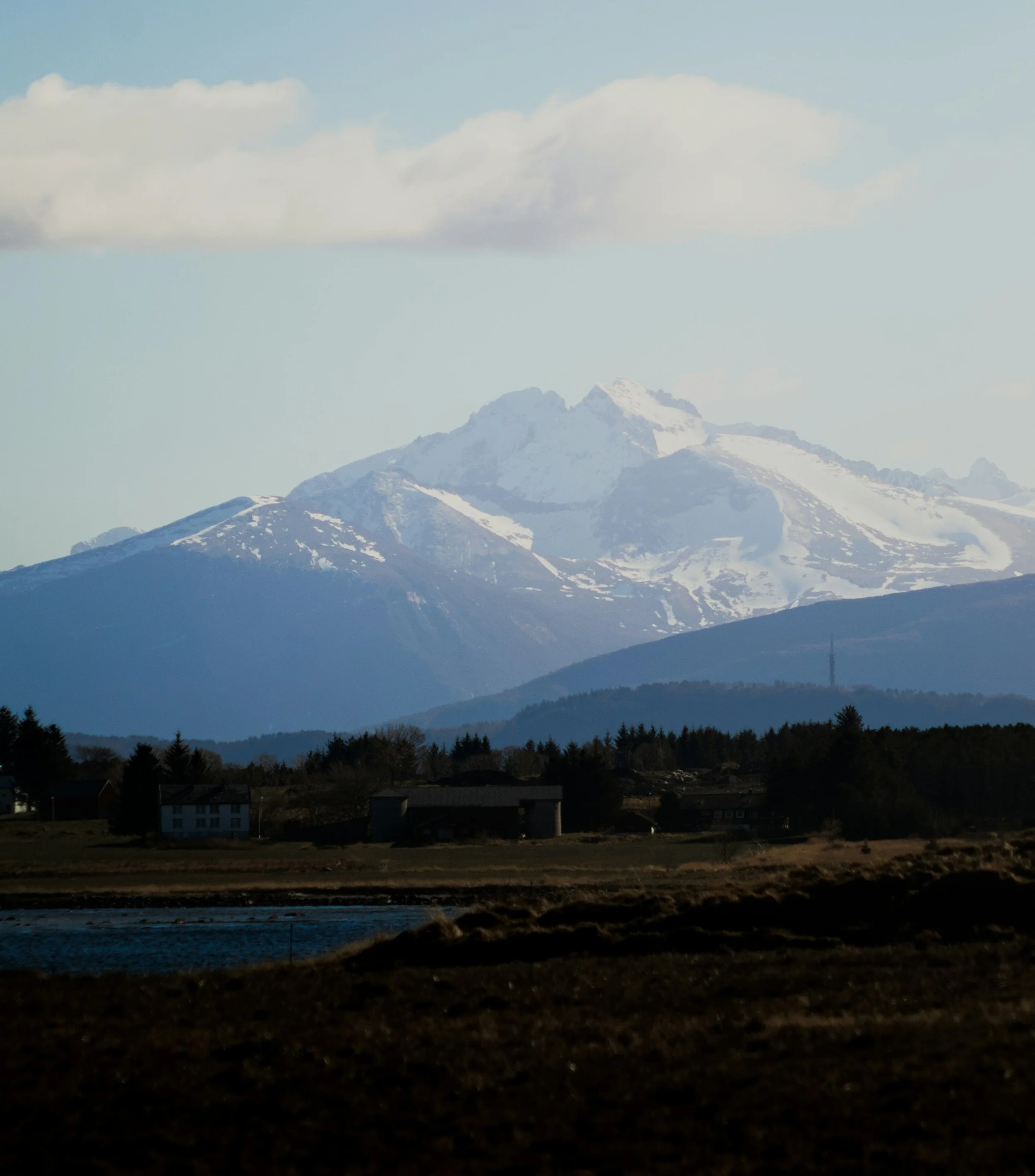 Snow-capped mountains in the distance with a partly cloudy sky, dark trees, and houses in the foreground near a body of water.