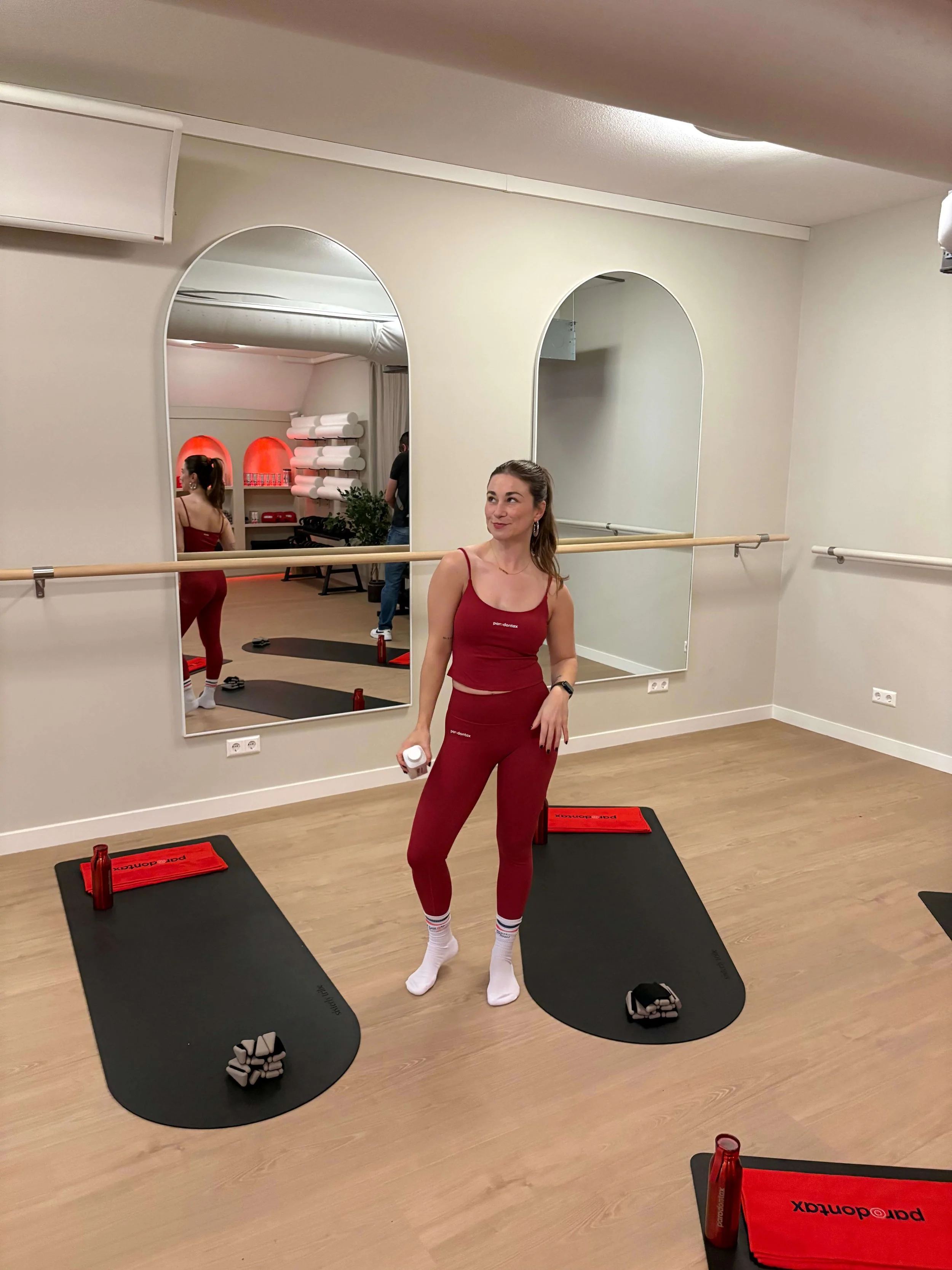Een vrouw in rode sportkleding staat in een fitnessruimte voor een spiegel, met sportmatten en gewichtjes op de vloer, en houdt een kleine witte bidon in haar hand.
