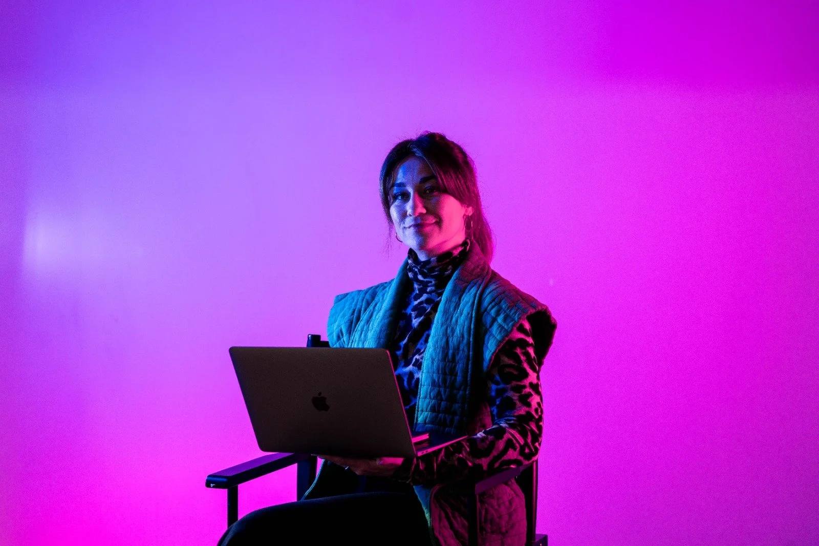 Vrouw zit op een stoel met een laptop, kleurrijke neon verlichting in de achtergrond.