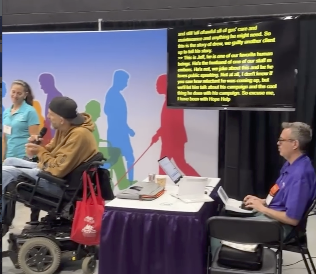 A captioner provides real-time captions during a live community event while a speaker using a power wheelchair addresses the audience.