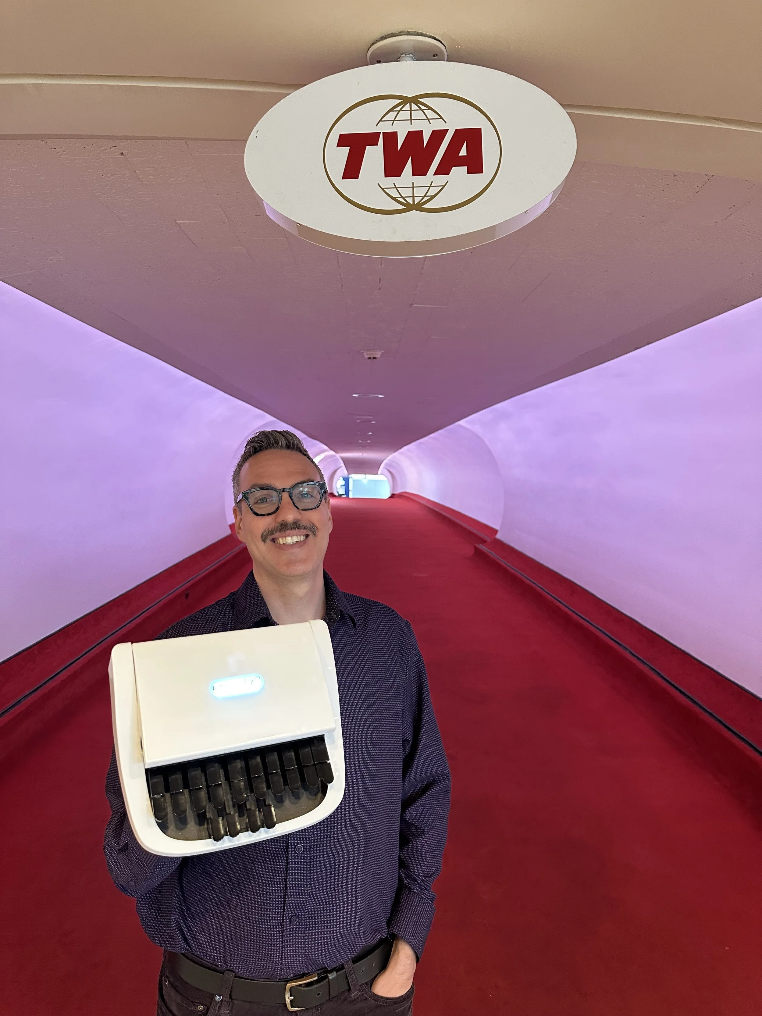 Thomas Herman holding a steno machine in the TWA Hotel jet bridge, smiling beneath the iconic TWA logo.