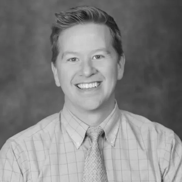 Christopher smiling at the camera he is a white man with short hair wearing a plaid shirt and tie