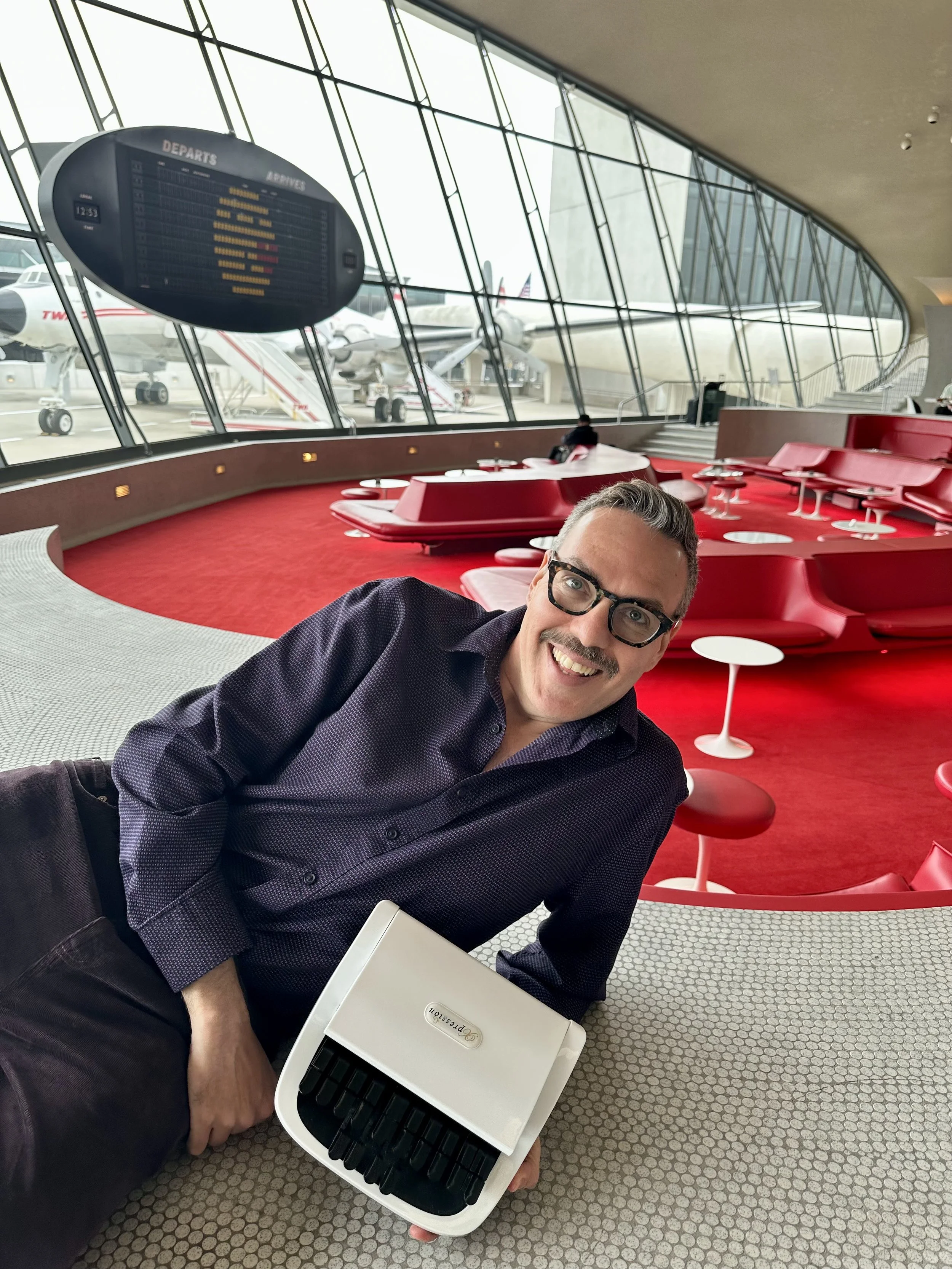 Thomas Herman smiling and seated on the floor of the TWA Hotel flight center at JFK, holding a Precision steno machine, with the iconic red lounge and vintage departure board visible behind him.