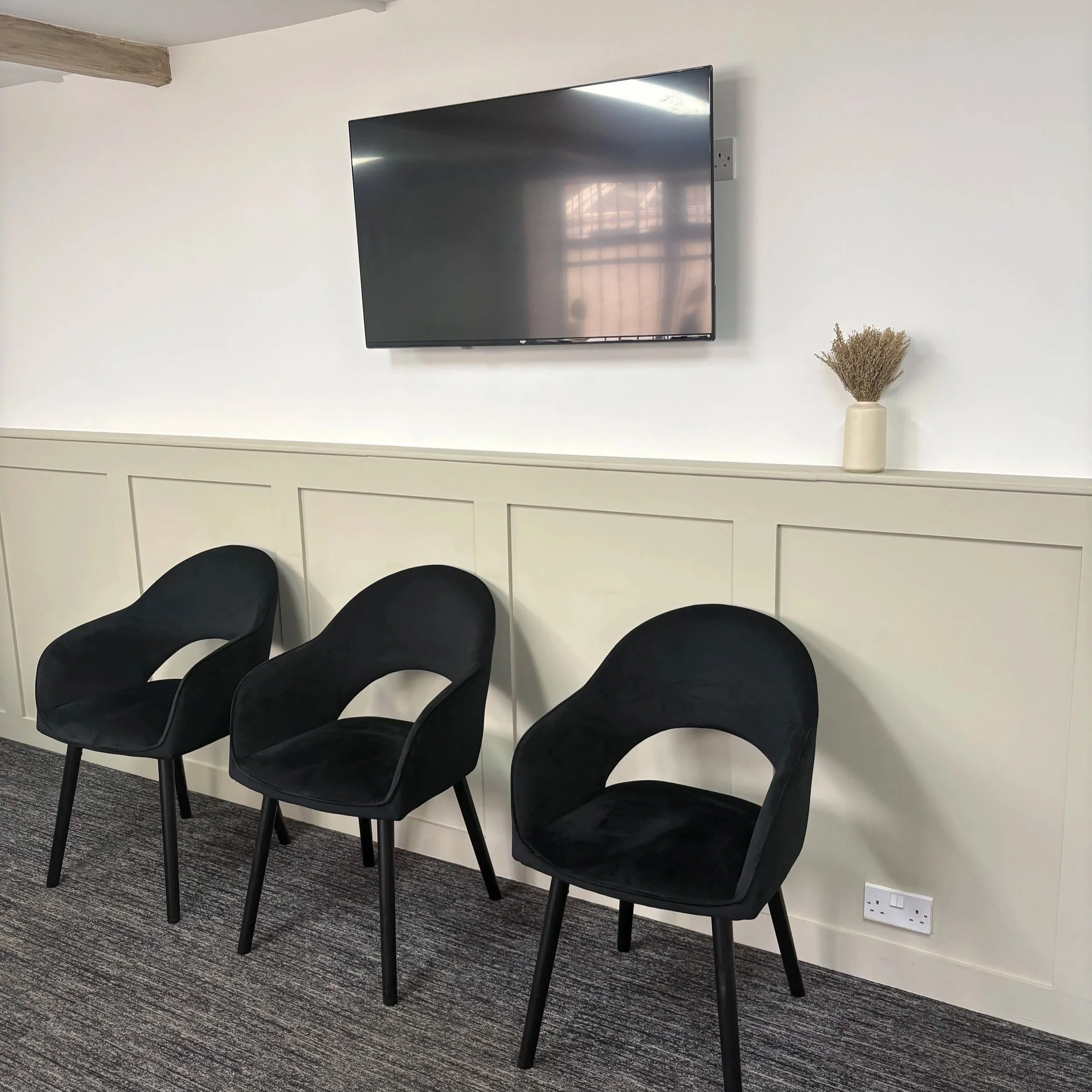 Four black chairs with curved backs and armrests are arranged along a white wall in a room with a dark gray carpeted floor. On the wall, a large flat-screen television is mounted above a white cabinet. To the right of the chairs, a beige vase filled with dried flowers sits on the cabinet. A white electrical outlet is visible on the wall near the floor.