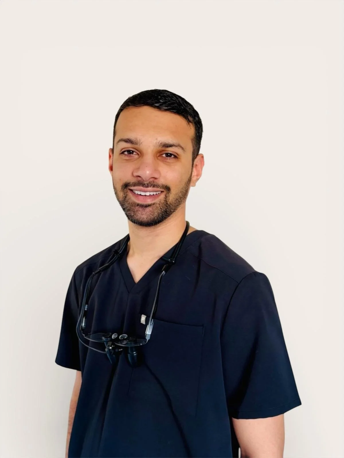 Dr Amish Patel. A smiling healthcare professional with short dark hair and a beard, wearing navy scrubs and a stethoscope around his neck, standing against a plain white background.