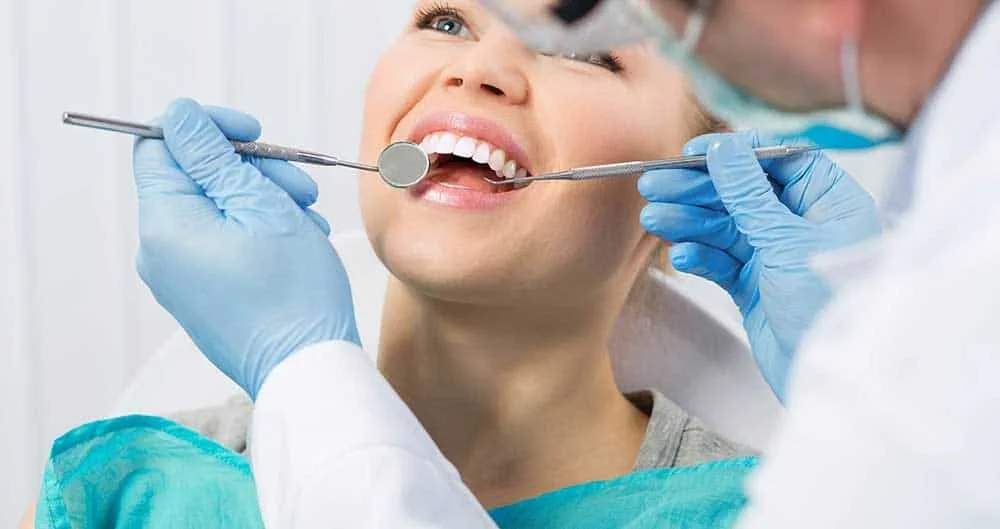 A woman at the dentist's office having her teeth checked with dental tools held by a dentist wearing gloves