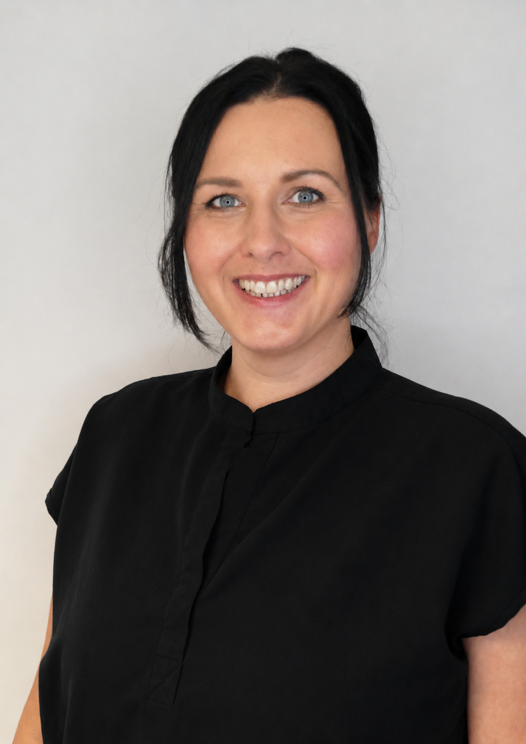 Portrait of a woman with short black hair, blue eyes, and a black top, smiling and standing against a plain, light-colored background.