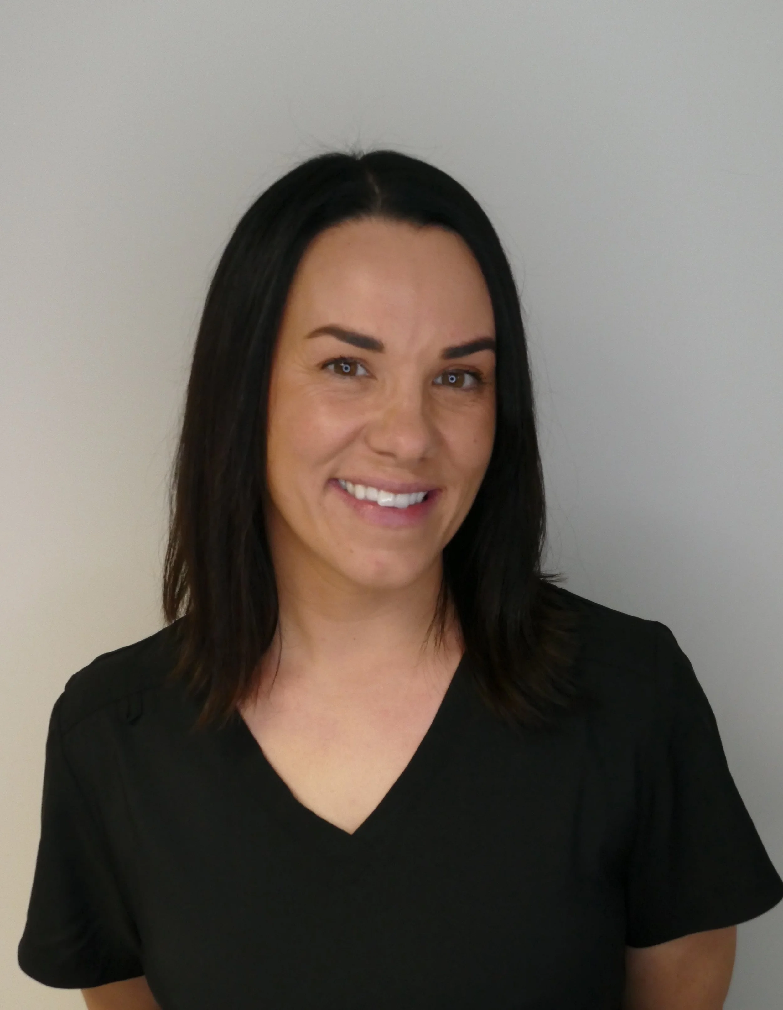 A woman with black hair and light skin, wearing a black V-neck shirt, smiling, standing in front of a plain light-colored wall.