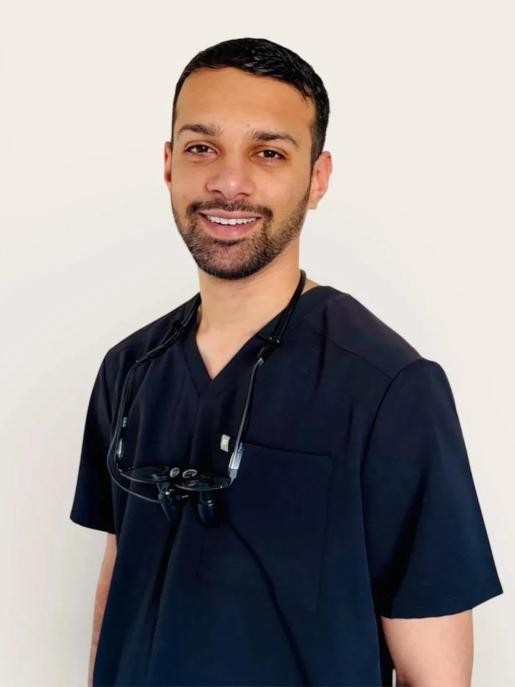 A smiling male healthcare professional wearing black scrubs and a stethoscope around his neck, standing against a plain white background.
