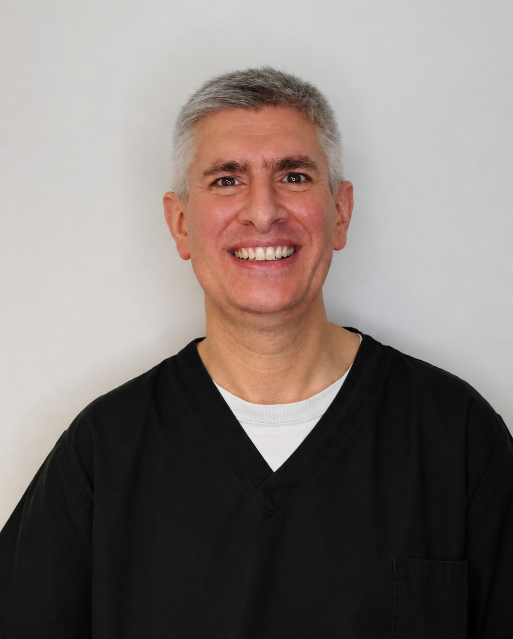 A smiling man with short gray hair, wearing a white shirt underneath a black medical scrub top, posing against a plain light-colored background.