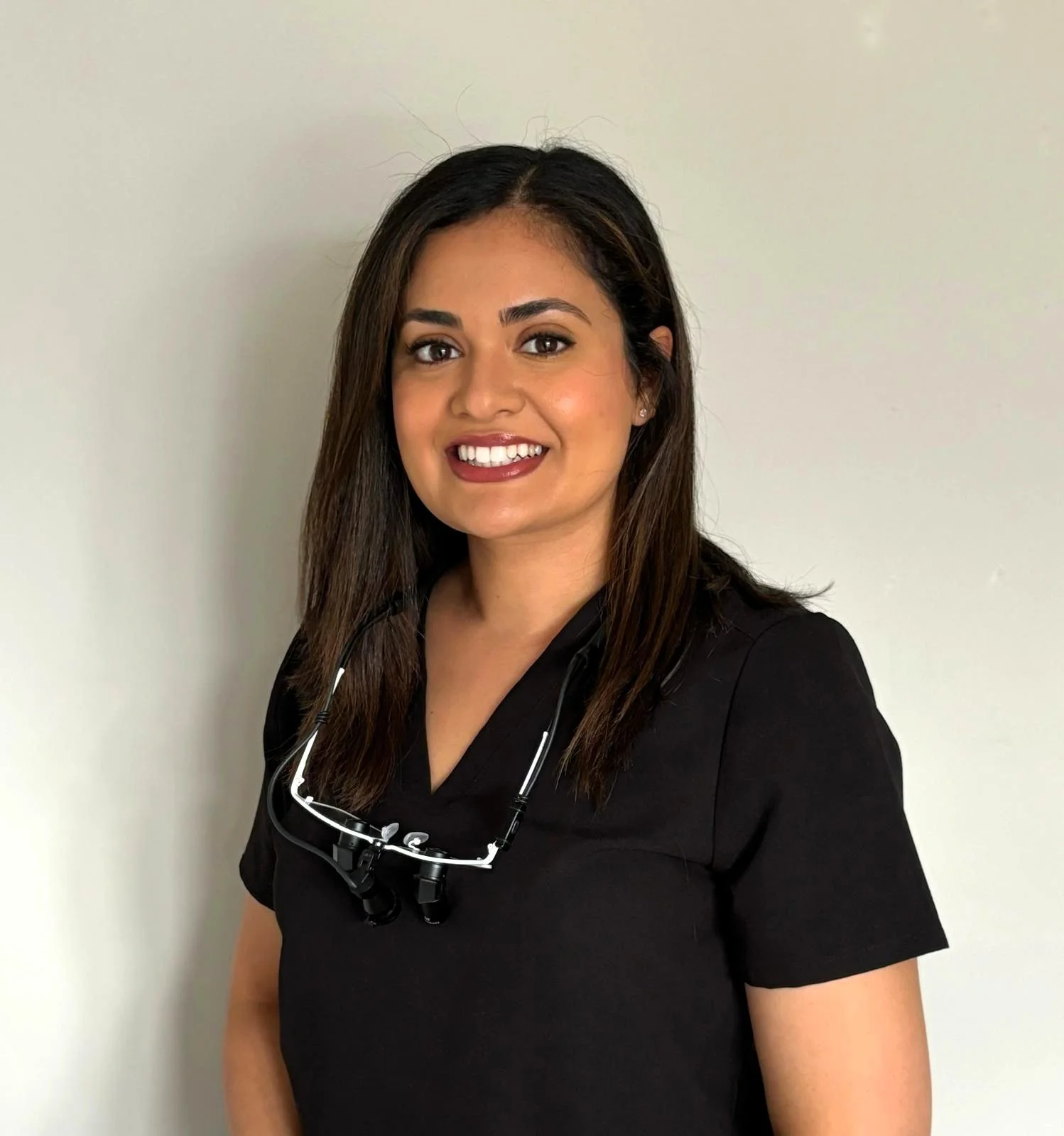 A smiling woman with dark hair, wearing black scrubs and a stethoscope around her neck, standing against a plain light-colored wall.