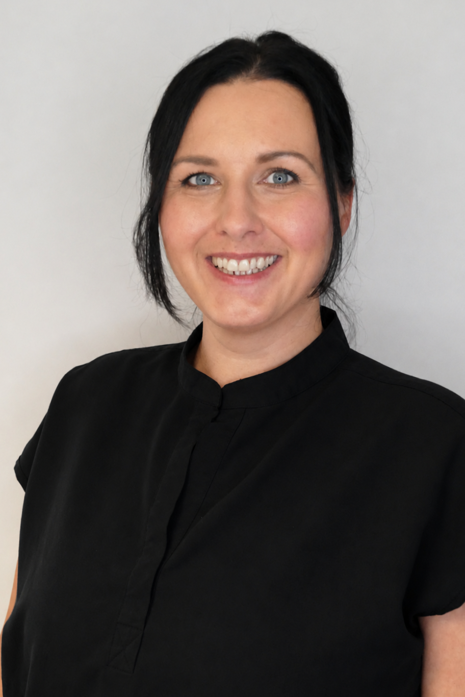 A woman with black hair and blue eyes smiling, wearing a black high-neck blouse, standing against a plain white background.