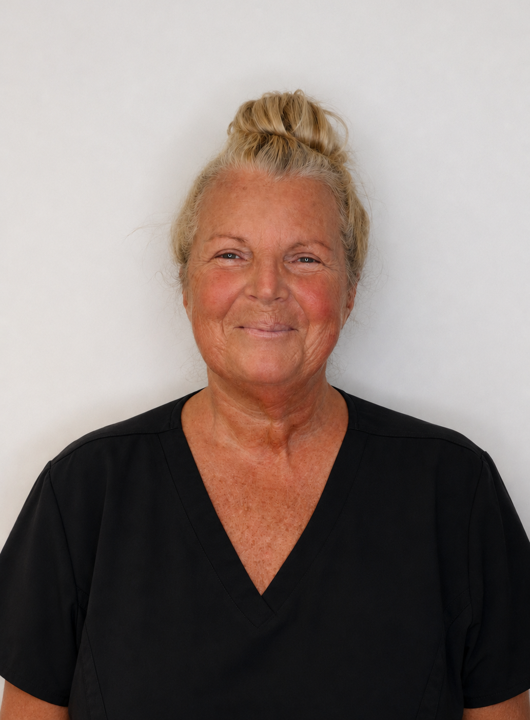 Portrait of an elderly woman with blonde hair tied up in a bun, wearing a black medical scrub top, smiling softly against a plain white background.