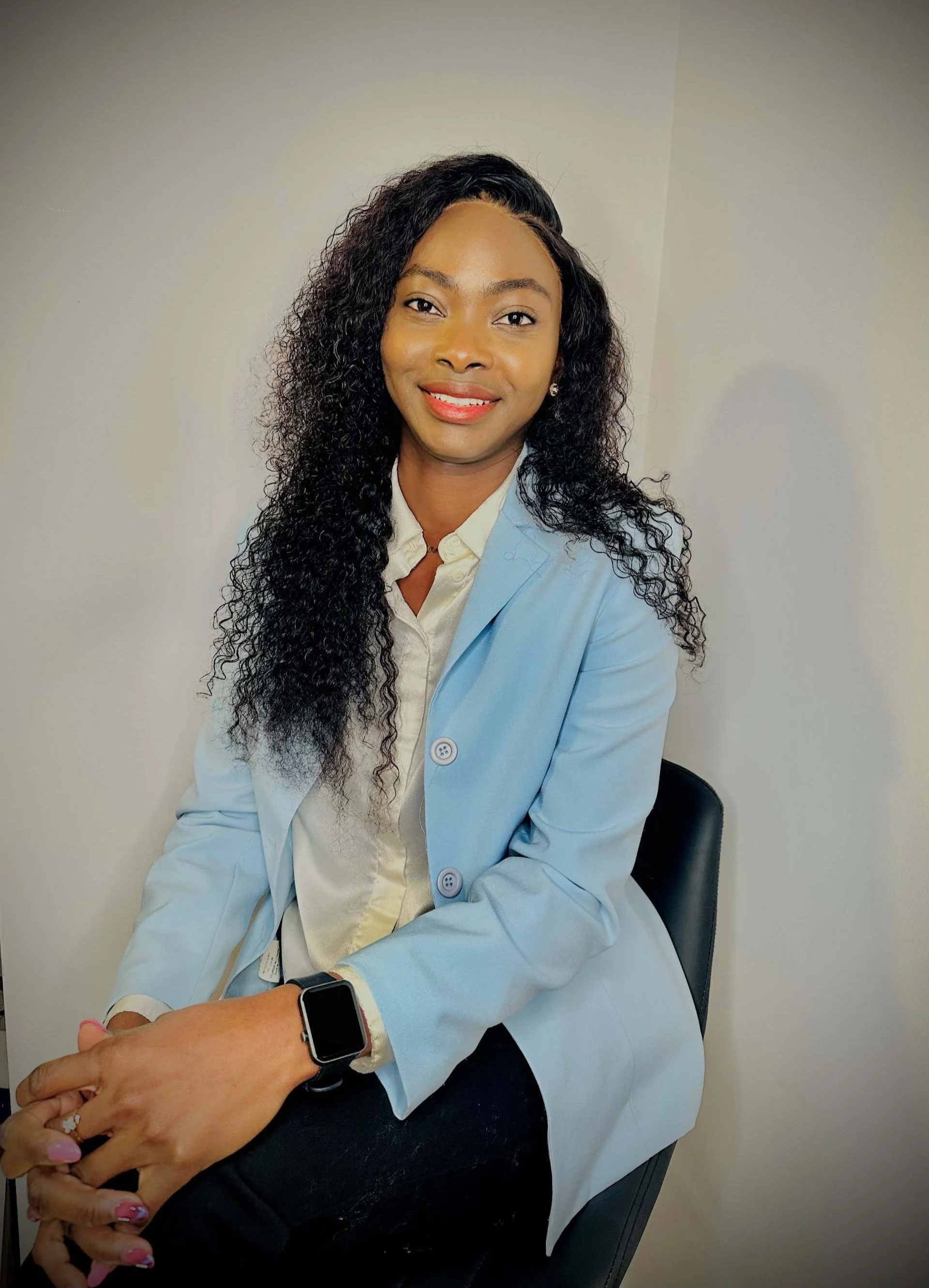 Femme afro-américaine portant un blazer bleu clair et une montre connectée, assise sur une chaise, souriant, dans un environnement intérieur