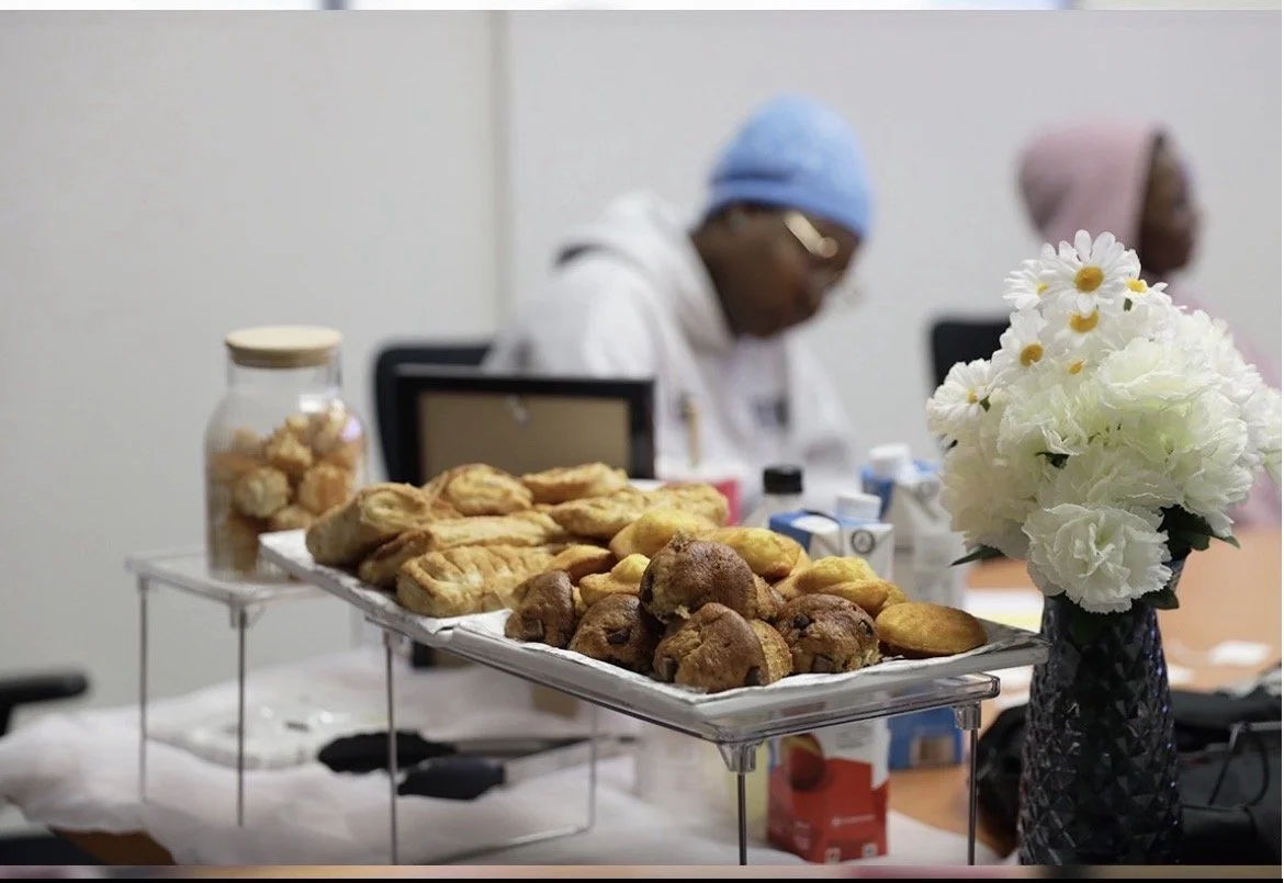 Plate de petits fours et muffins sur une table, avec un vase de fleurs blanches, dans une salle de réunion occupée par deux personnes en arrière-plan.