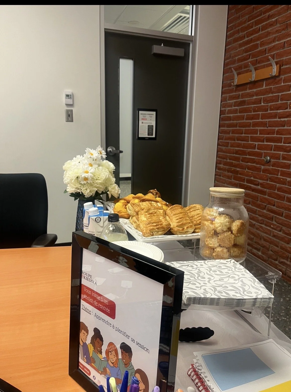 Table avec des viennoiseries, une jarre de cookies, une vase avec des fleurs blanches, un cadre avec un affichage, et des documents, dans une salle de réunion ou un espace de travail.