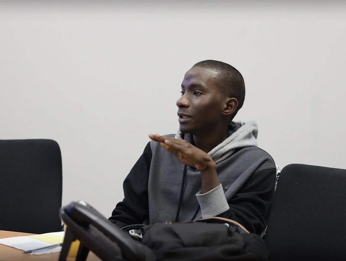 Un jeune homme assis à une table lors d'une réunion, portant une veste à capuche grise et un t-shirt noir, gesticulant avec la main lors de la discussion.
