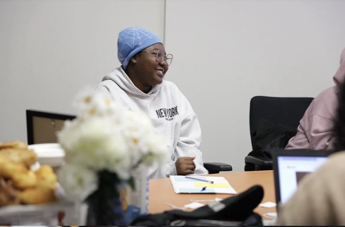 Une femme souriante portant un sweat blanc avec l'inscription «New York» et un chapeau bleu, assise à une table lors d'une réunion, avec des documents et une fleur en premier plan.
