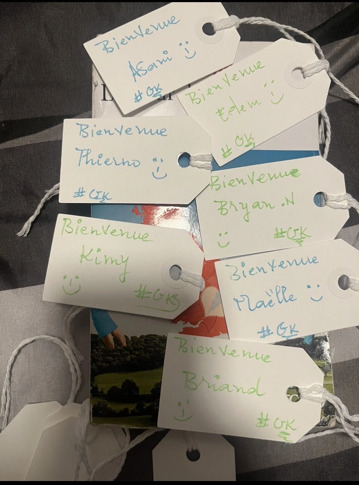 Groupe de plusieurs étiquettes avec des messages de bienvenue écrits en différentes couleurs. Chaque étiquette a un nom, un smiley et un hashtag '#GK'. Certaines étiquettes portent les noms de Asavi, Edem, Thierno, Bryan.N, Kimy, Maëlle, Brand. L'ens