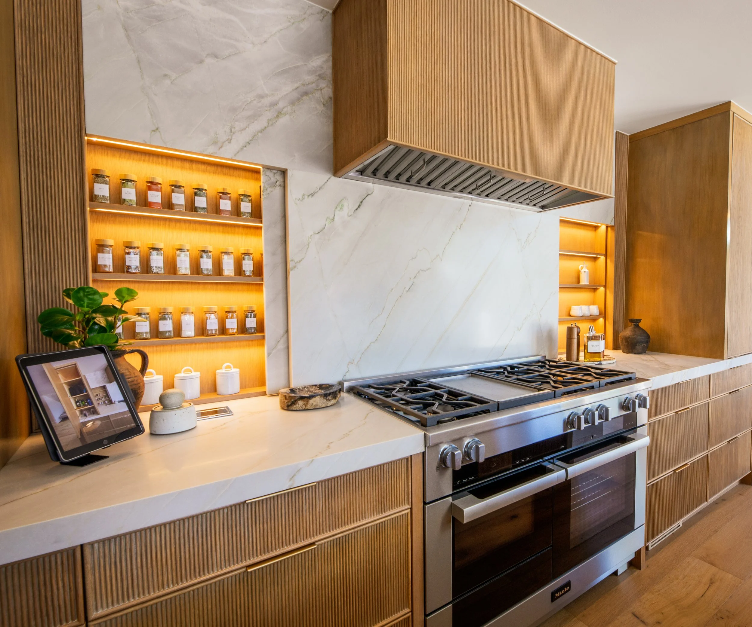 Modern kitchen with wooden cabinetry, marble backsplash, and stainless steel gas stove. Display shelves with jars and decorative items, and a tablet on the countertop.
