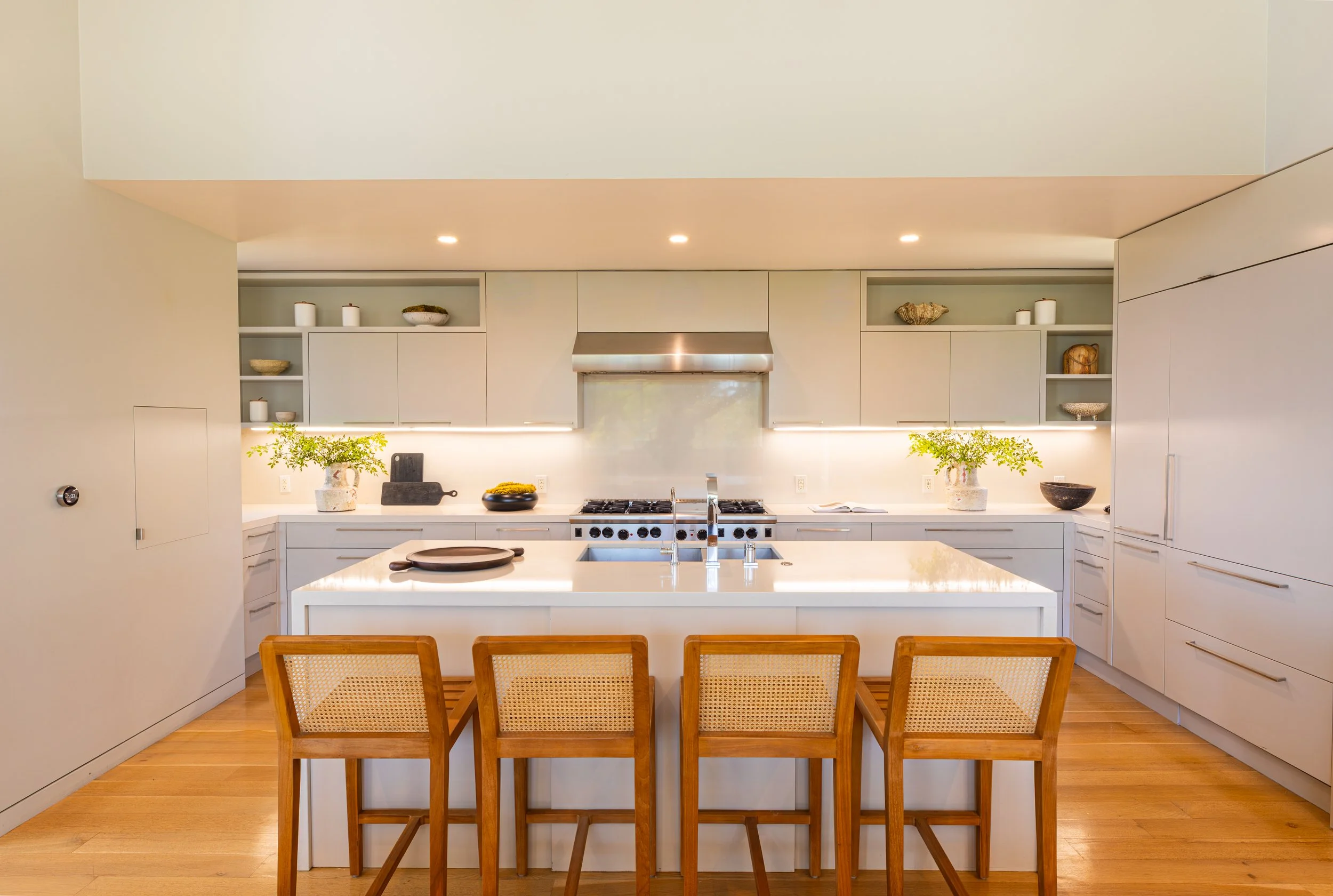 Modern kitchen with white cabinetry, a large island with a white countertop, and wooden dining chairs. Decorative bowls and plants are on the counter, with built-in shelves and a stainless steel range hood.