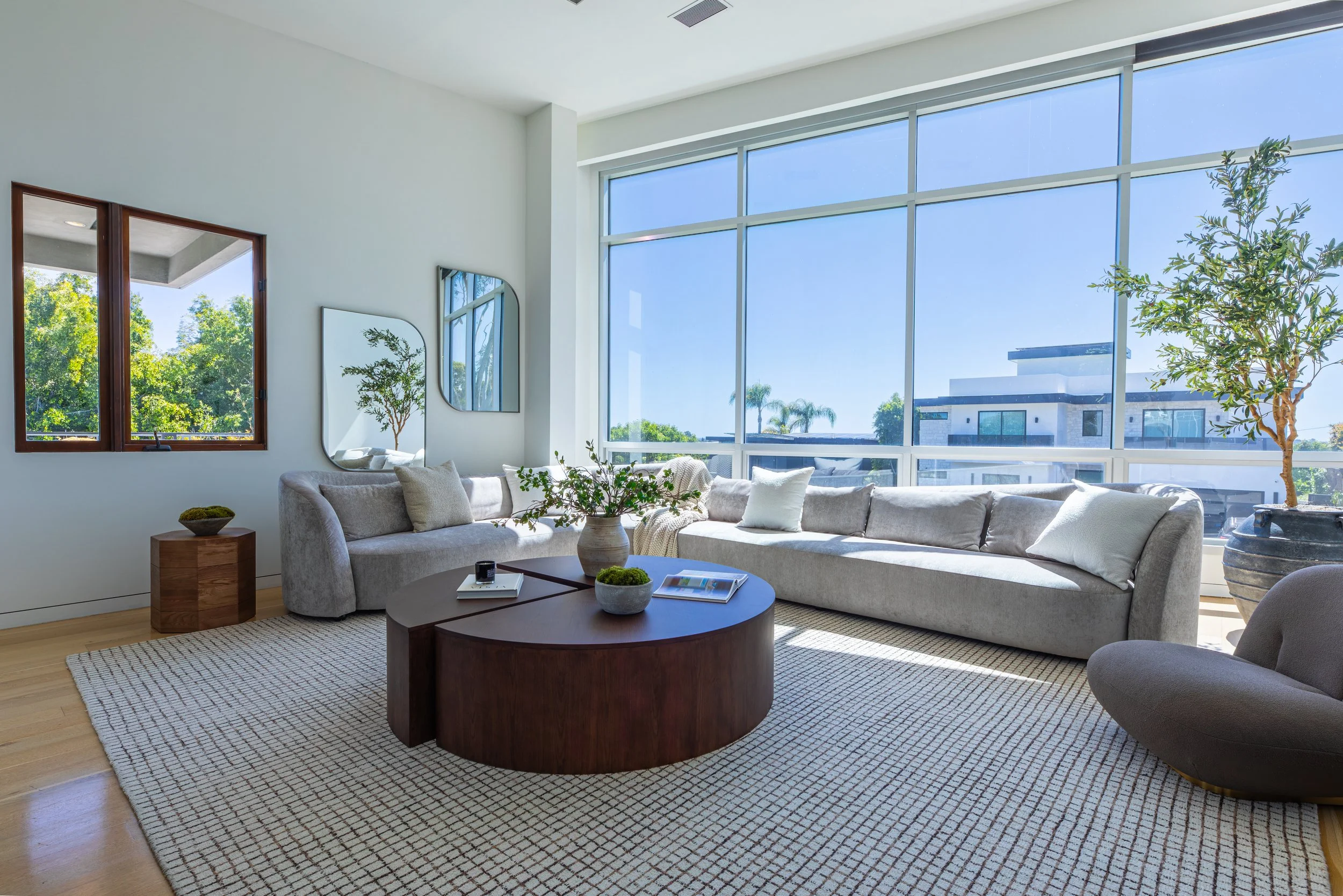 Bright living room with large windows, a sectional sofa with pillows, a round wooden coffee table with books and plants, a small side table, and wall mirrors, with greenery visible outdoors.