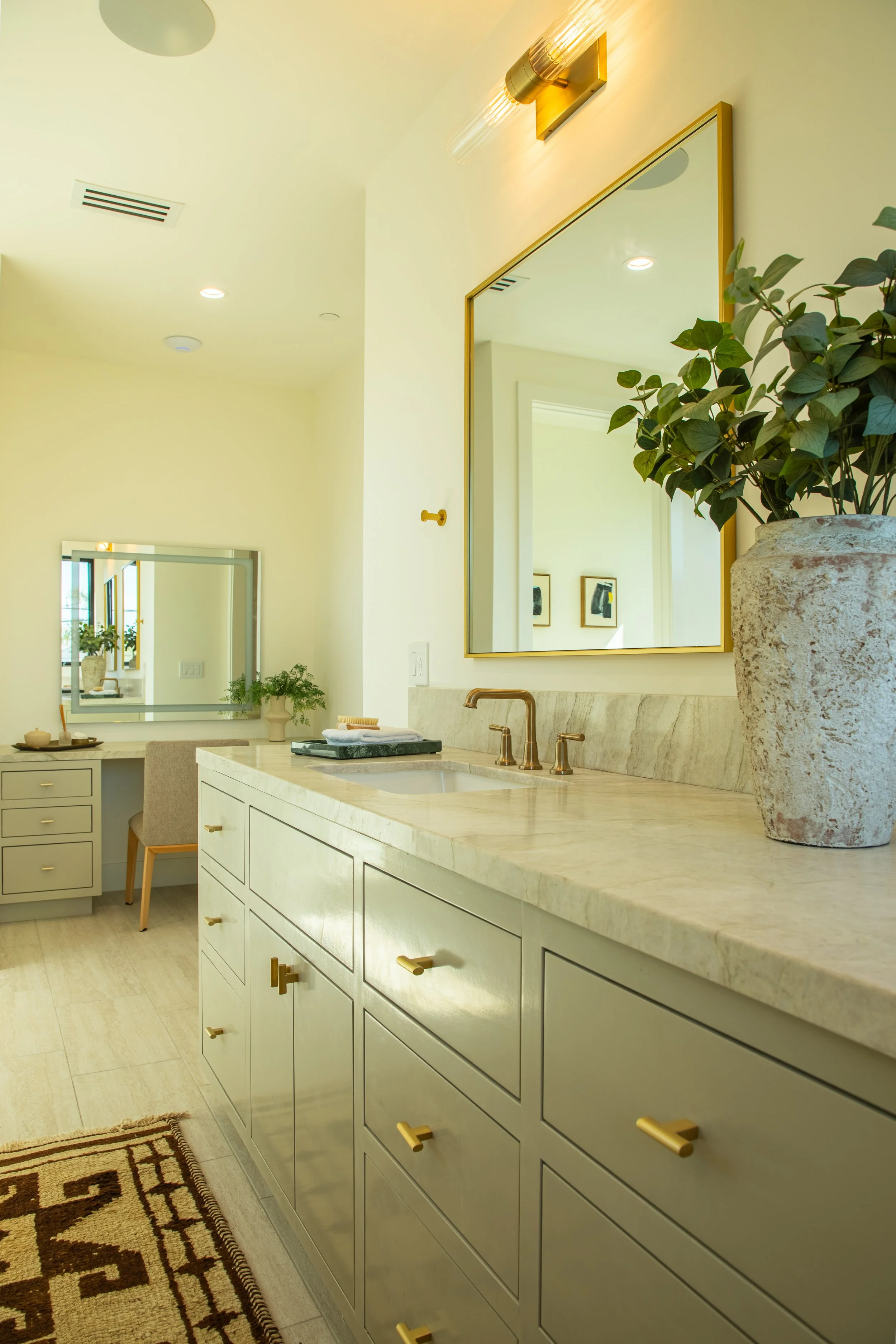 A bathroom with light green cabinetry, a marble countertop, and a large mirror with a gold frame. There is a potted plant on the counter and a patterned rug on the floor.