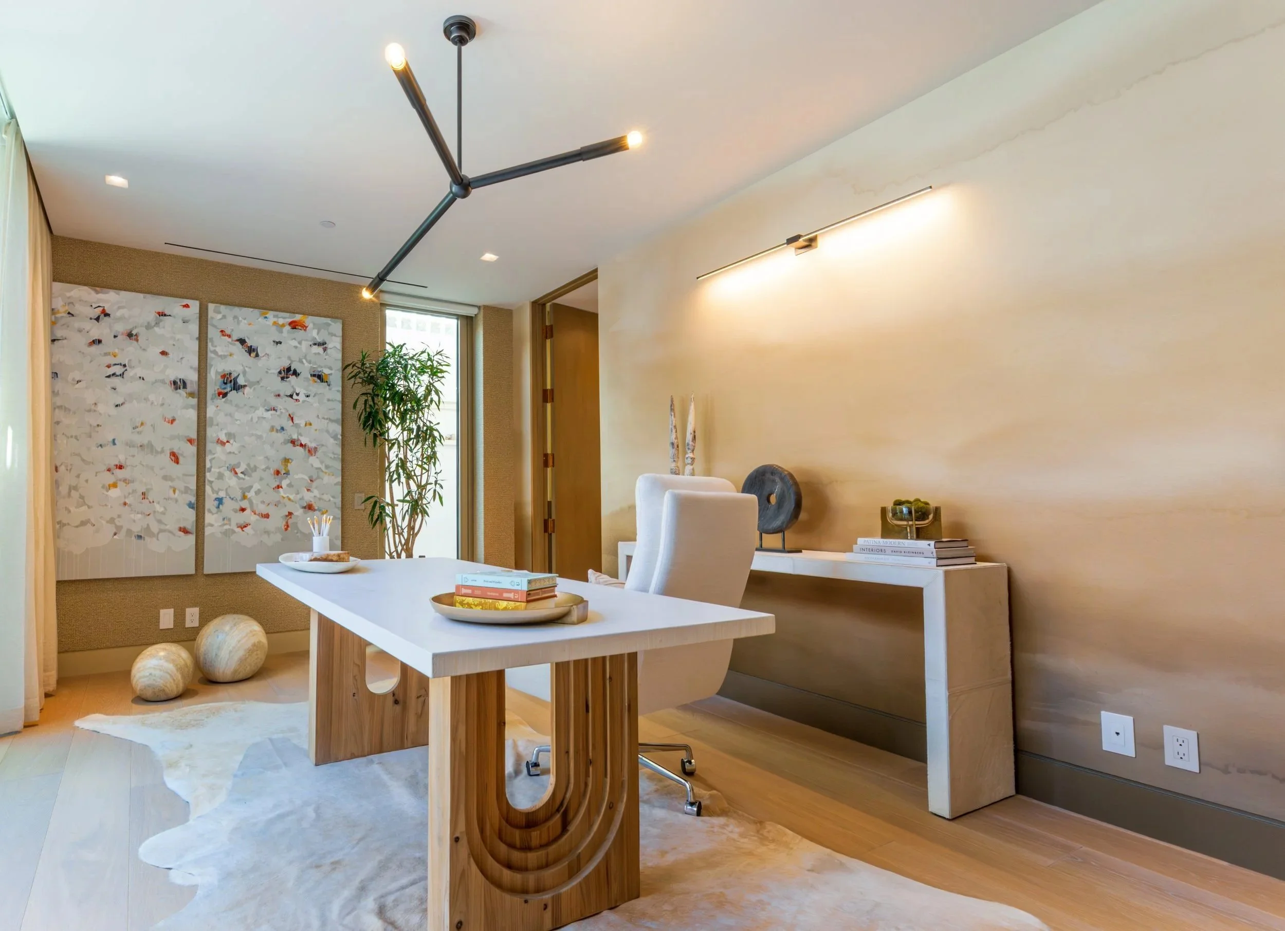 A modern dining and living room area with a white table, beige chairs, green plant by the window, abstract artwork on the wall, and contemporary lighting fixtures.