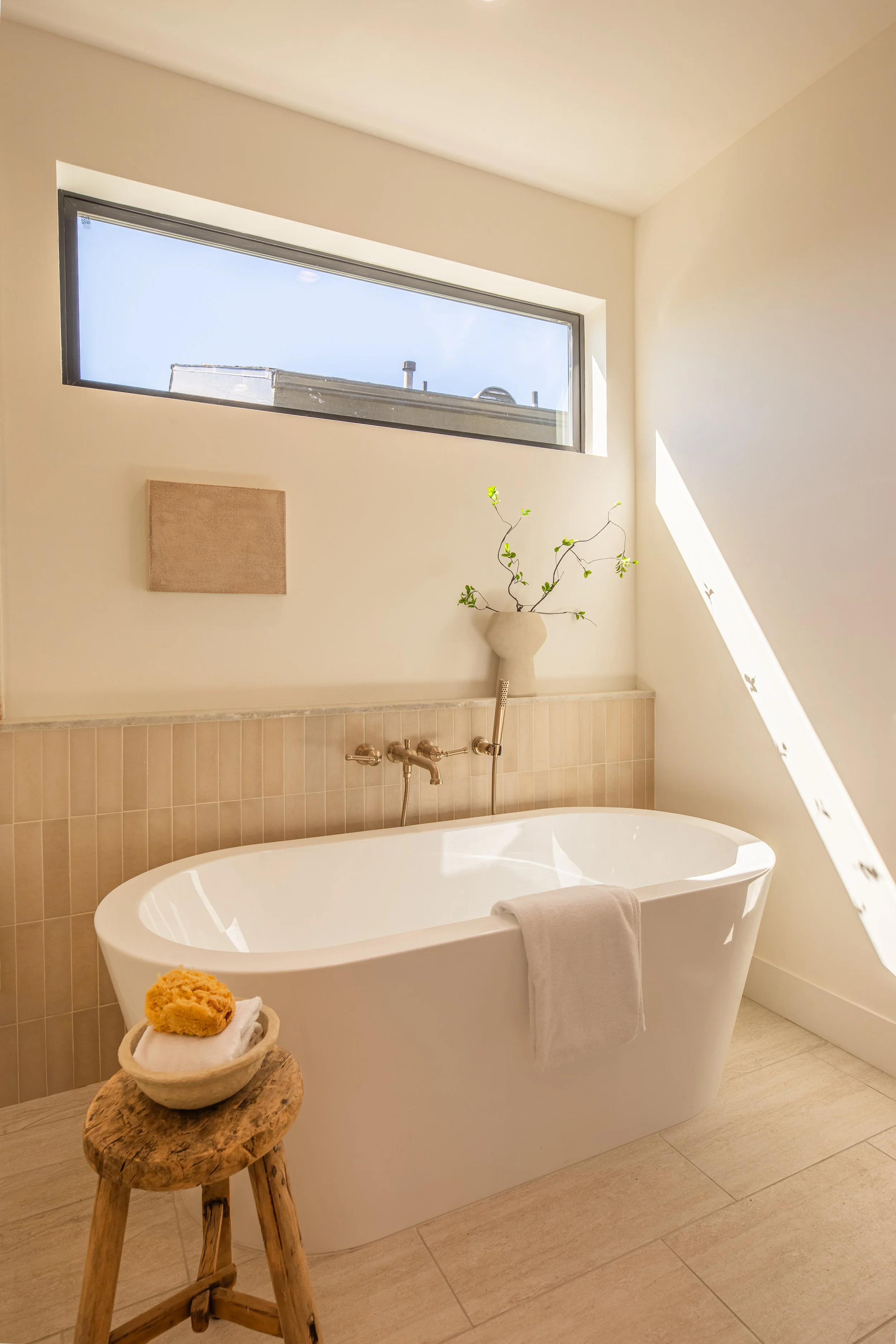 A modern bathroom with a free-standing bathtub, a small wooden stool with towels and a sponge, a potted plant on a pedestal, a rectangular window letting in natural light, and beige tiled walls and floor.