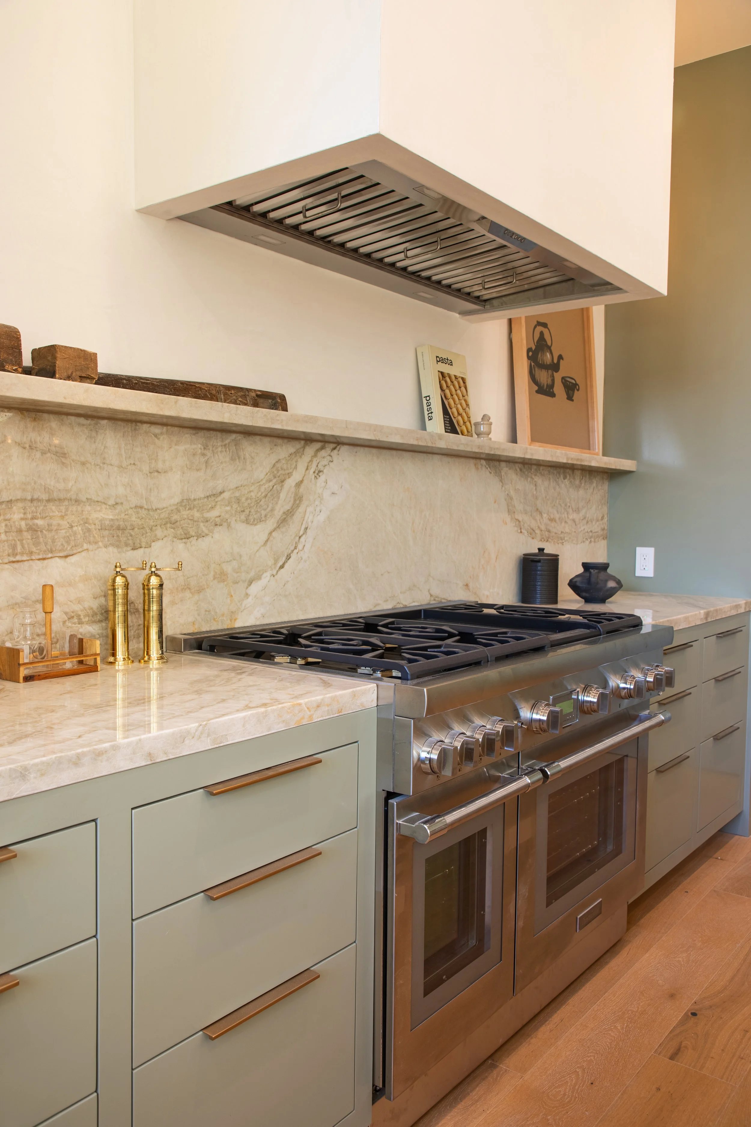 A modern kitchen with a marble countertop, a stainless steel gas stove, beige cabinets with copper handles, a marble backsplash, a coffee maker, and decorative items like black and beige vases. There is also a framed picture and a wooden tray on the counter.