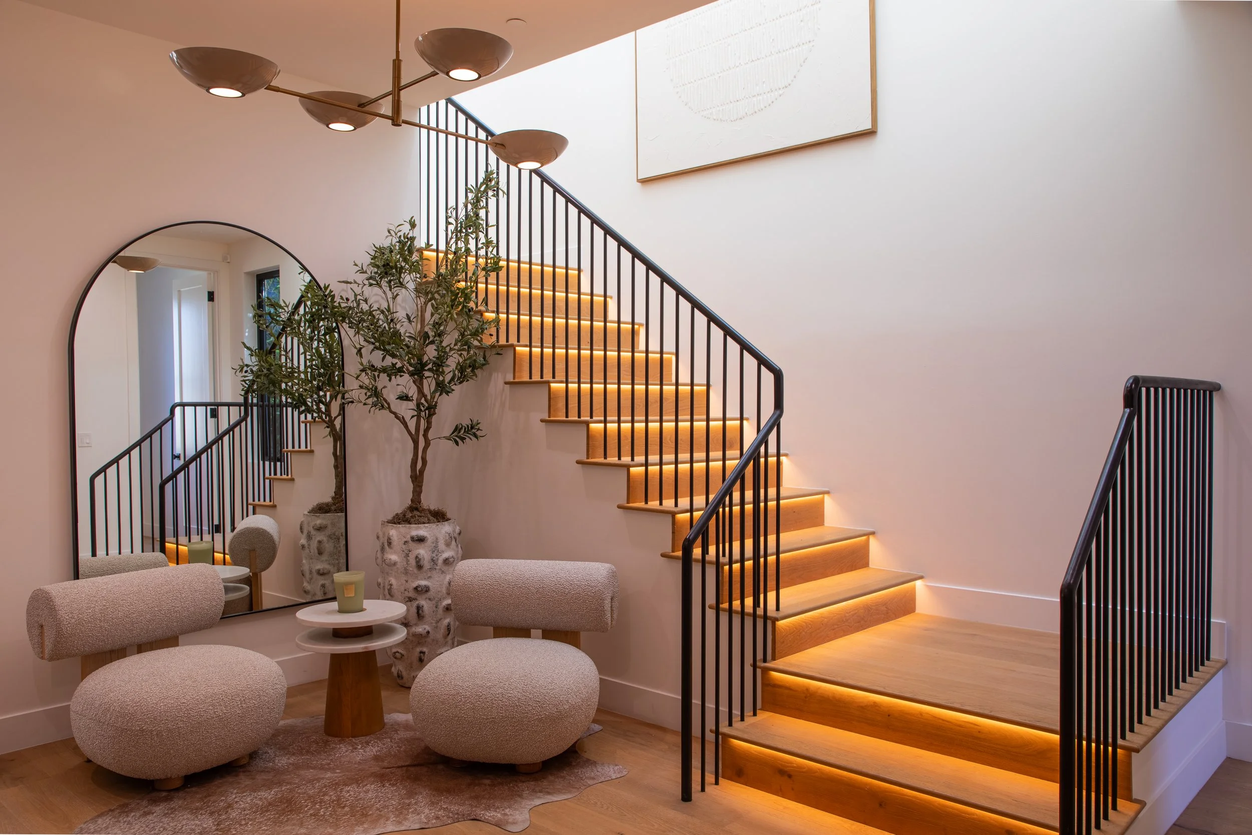 Interior view of a modern staircase with wooden steps and LED lighting, residing beside a seating area with beige rounded chairs, a small round table, tall potted plants, and minimalist decor including a large round mirror and abstract artwork.