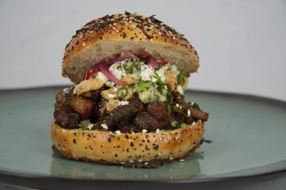 A sandwich with shredded meat, chopped tomatoes, pickled red onions, fresh cilantro, and crumbled white cheese in a sesame seed bun.