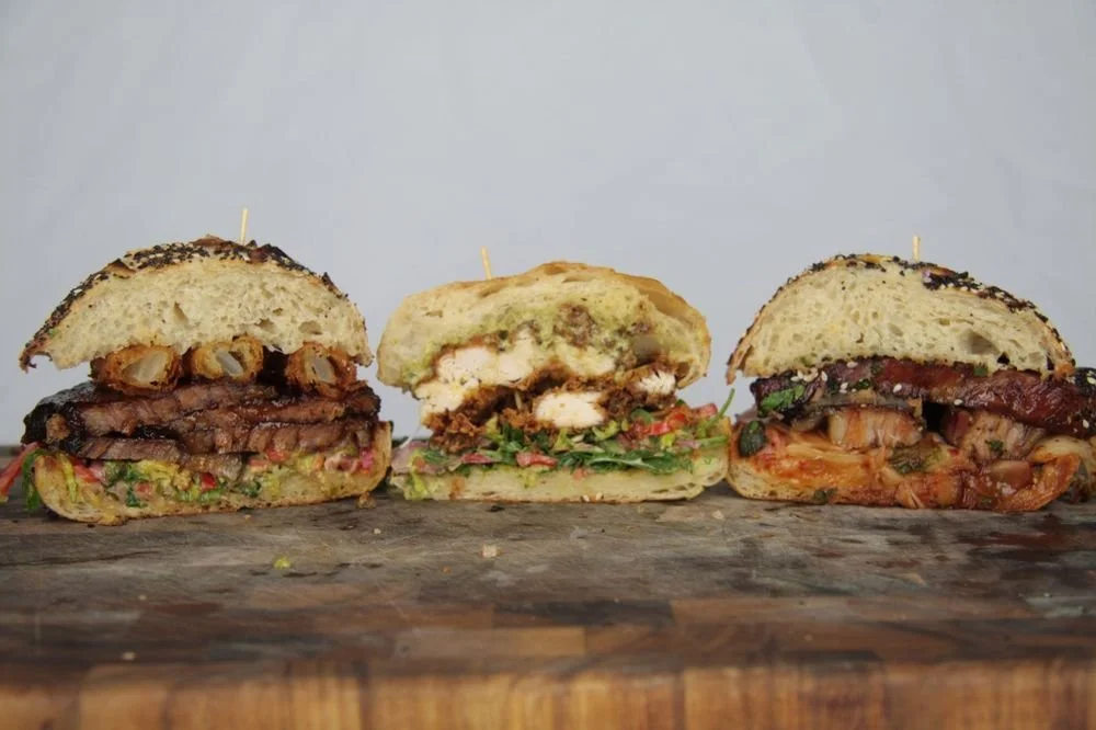 Three different sandwiches on a wooden surface, with various meats, vegetables, and bread.