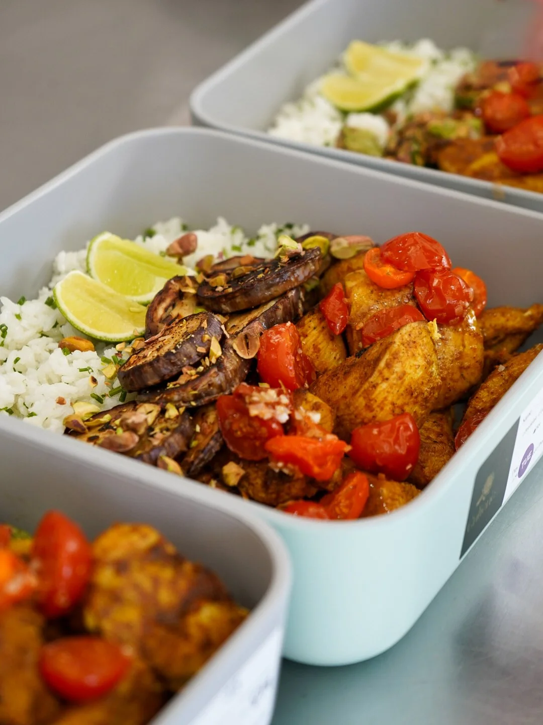 Meal prep containers with rice, grilled vegetables, and chicken, garnished with lime wedges.