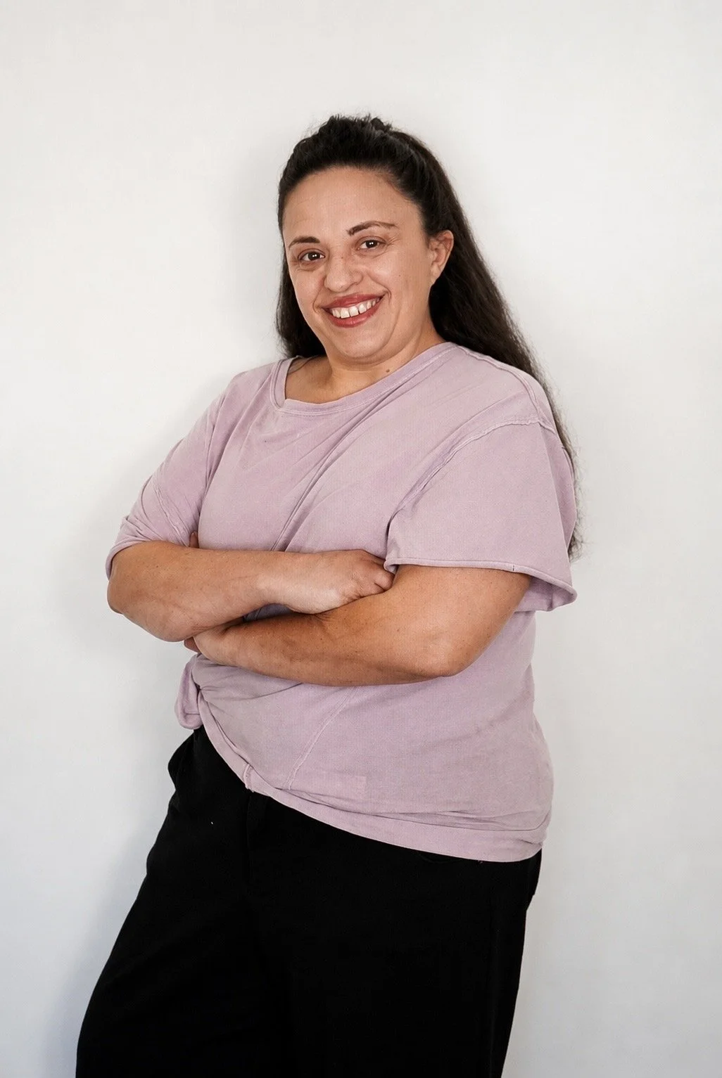 A woman with long dark hair standing against a plain white wall, wearing a light purple shirt and black pants, smiling with arms crossed.
