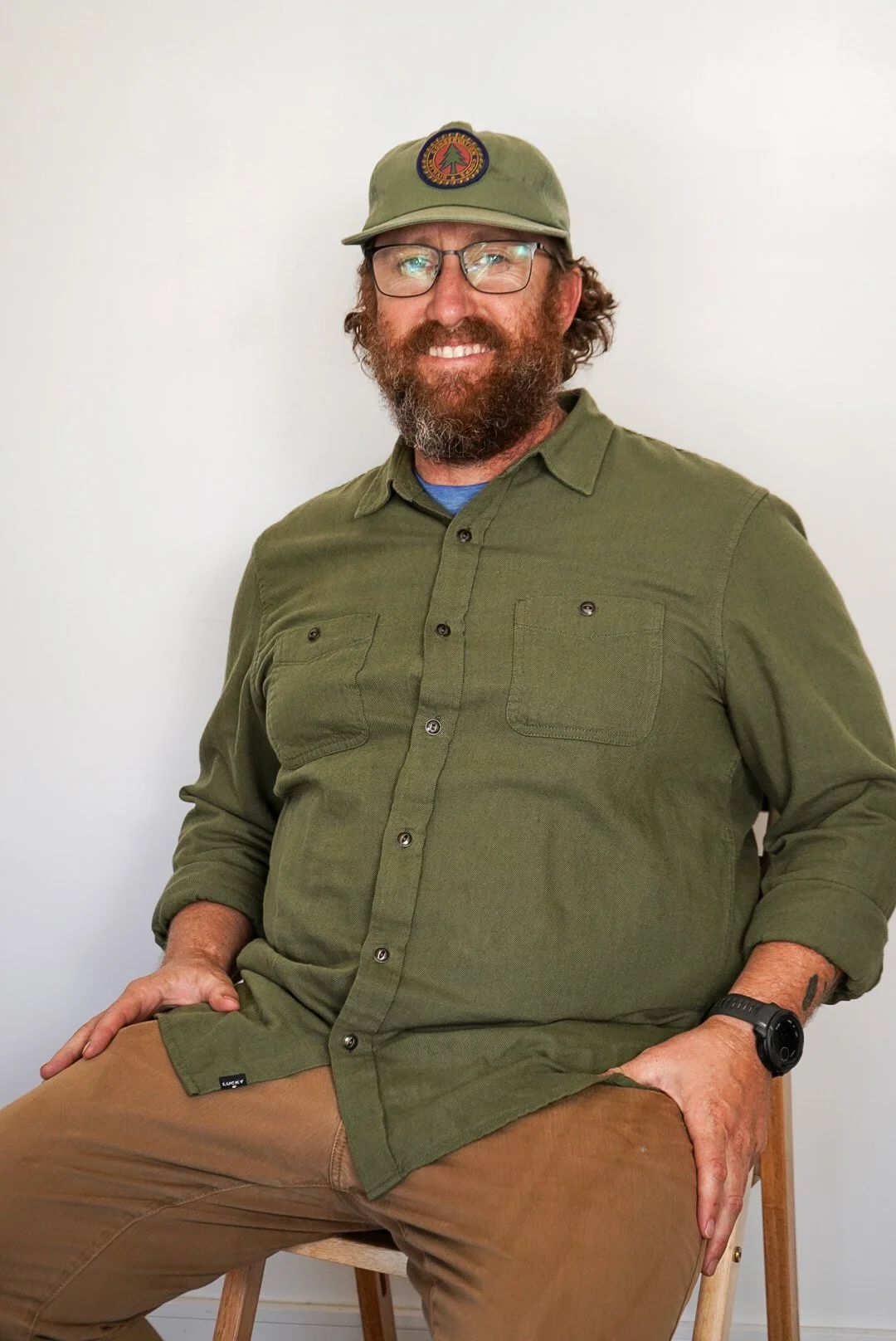 A man with glasses, a full beard, and a hat sitting on a wooden chair against a plain white wall. He is wearing an olive green button-up shirt, khaki pants, and a wristwatch.